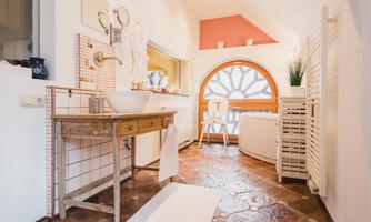 A bright and inviting bathroom with an elegant bathtub and a round window. The floor is made of rustic stone and a wooden table complements the cozy atmosphere.