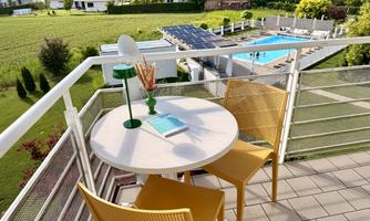 A cozy balcony with a round table and two yellow chairs. In the background, there is a pool and a green landscape.