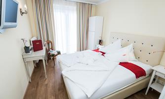 A bright hotel room with a double bed and modern furniture. A television hangs on the wall, and large windows provide plenty of daylight.