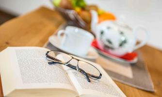 An open book lies on a wooden table, next to it are a pair of glasses and a cup with a saucer. In the background, you can see a teapot and a bowl with fresh fruit.