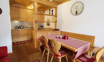 A cozy kitchen with wooden furniture and a large dining table. On the table is a teapot, and the walls are adorned with decorative elements.