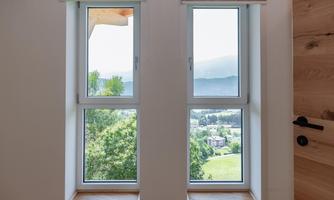 A light-flooded room with two windows overlooking a green landscape. In the background, mountains and trees are visible.