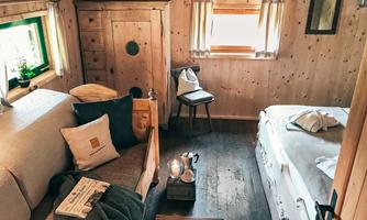 A cozy room with wooden walls and two beds. On a table, there are a few drinks and a newspaper.