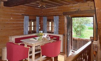 A cozy room in a wooden house with a table and red chairs. Through the open door, you can see the greenery.