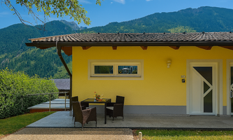 A modern, yellow house with a porch. In the background, green mountains stretch under a blue sky.