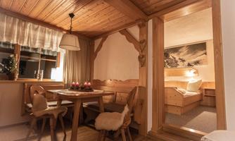 A cozy wooden room with a dining table and chairs. In the background, there is a bedroom with a pleasant, warm light.