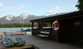 A wooden terrace by the shore of a lake, with a view of high mountains. Two toy-like fish are lying next to a bench and a life ring.