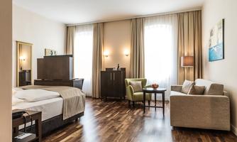 A modern hotel room with a double bed, a cozy sofa, and a small table. Large windows bring plenty of light into the room.