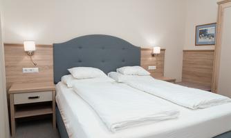 A modern bedroom with two single beds and a padded headboard. The walls are light, and there are nightstands with lamps.