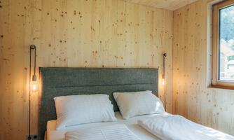 A modern bedroom with wooden walls and a cozy double bed. Two pendant lamps illuminate the room, and a window lets in plenty of daylight.