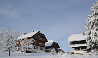 A winter landscape with snow-covered houses and trees. The sky is clear and blue.