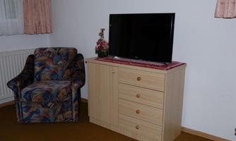 A living room with a television on a dresser and a comfortable armchair. The walls are light, and there are curtains at the windows.
