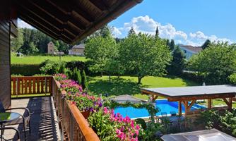 A balcony with blooming plants and a view of a pool. In the background, green meadows and trees can be seen.