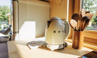 A stylish kettle from SMEG stands on a wooden table. Next to it, cooking spoons are visible in a container and a refrigerator is seen in the background.