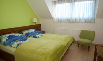 A cozy bedroom with a large bed and colorful pillows. The walls are painted green, and there is a window with curtains.