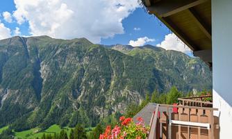 An impressive mountain landscape with green meadows and high peaks. In the foreground, a balcony with blooming plants and a radiant blue sky.