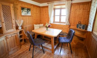 A cozy wooden room with a large table and four chairs. A flower vase stands on the table, and light comes through a window.