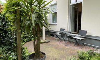 A cozy outdoor area with a small table and chairs. Surrounded by plants and green spaces.