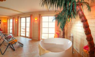 A relaxing wellness area with a round bathtub and a palm tree. The room has large windows and warm light for a cozy atmosphere.