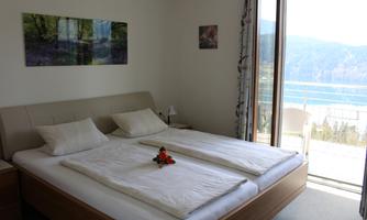 A bright bedroom with a double bed and two pillows. The windows offer a view of the mountains and a balcony.