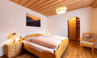 A cozy bedroom with a large double bed and wooden furniture. A picture of a landscape hangs on the wall, and there is a nice chair in the corner.