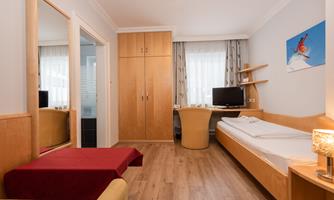 A modern room with wooden furniture and a single bed. In the background, a television and a large mirror can be seen.