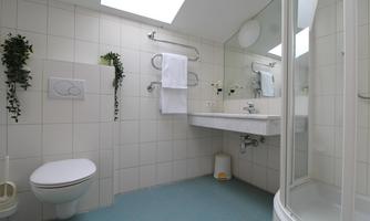 A modern bathroom with white tiles and a large mirror. There is a toilet area, a shower, and fresh towels.