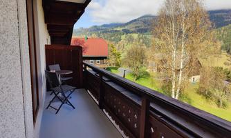 A balcony with two chairs and a table. In the background, green hills and trees can be seen.