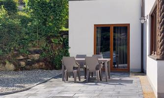 A cozy outdoor area with a table and chairs on a paved floor. In the background, there is a green garden and a wall with windows.