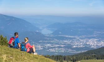 A couple is sitting on a meadow and enjoying the view of a valley. In the background, there are mountains and a clear sky.