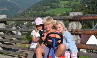 Three children are happily playing outdoors. They are sitting on a red toy tractor in front of a picturesque mountain landscape.
