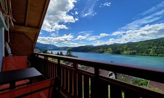View from a balcony overlooking a clear lake and green hills. The sky is blue with some clouds.