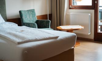A cozy hotel room with a bed, an armchair, and a small table. The view from the window shows an orange umbrella and plenty of light.