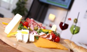 A beautiful wooden board with various types of cheese, ham, and fresh tomatoes. In the background, two colorful cocktails can be seen.