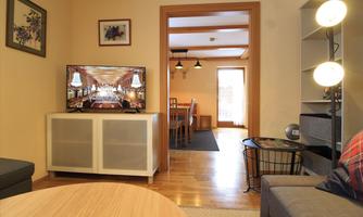 A cozy living space with a TV on a white cabinet. In the background, a dining table and lots of daylight can be seen.