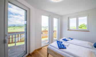 A bright bedroom with two single beds and large windows. The view overlooks a green landscape and a blue sky.