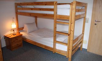 A bunk bed made of wood stands in a simple bedroom. Next to it is a nightstand with a lamp.