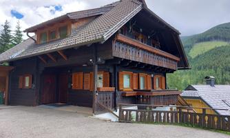 A charming wooden house in Alpine style with a green backdrop. The facade features large windows and a stylish balcony.
