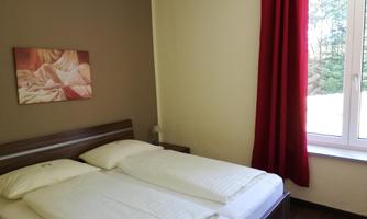 A bright bedroom with two single beds and a window that lets in daylight. A picture hangs on the wall, and the room has a warm color scheme.