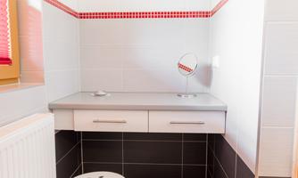 A bright bathroom with white and gray tiled walls. A small table and a stool with a large mirror and a towel are visible.
