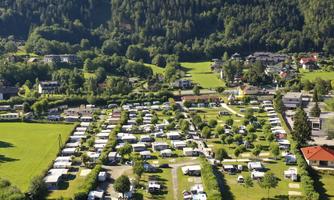A beautiful campsite by the water, surrounded by green meadows and hills. The pitches are neatly arranged and some boats are docked at the shore.