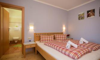A cozy bedroom with a double bed and checked pillows. In the background, a bathroom is visible.