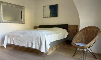 A bright bedroom with a large bed, a mirror, and a modern chair. The walls are plain and the decor is minimalist.