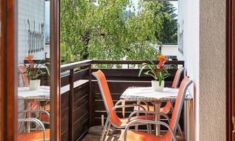 A sunny balcony with orange chairs and a table. In the background, green trees and a clear sky can be seen.