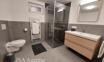 A modern bathroom with a shower, a wooden washbasin, and modern fixtures. The walls are kept in simple shades of gray.