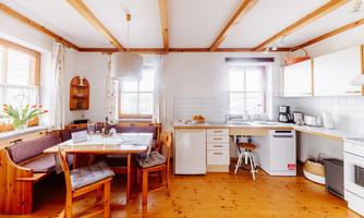 A bright kitchen with a wooden beam ceiling and windows that let in plenty of daylight. The dining area is cozy with a table and chairs, surrounded by a comfortable seating area.