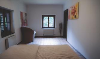 A simple and calm bedroom with a large window. Colorful pictures hang on the walls, and a comfortable armchair sits in the corner.