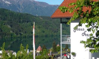 A picturesque landscape with mountains and a calm lake. In the foreground stands a building with a terrace, surrounded by trees and plants.