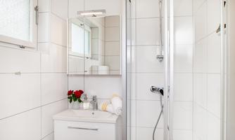 A modern bathroom with white tiles and a mirror. Fresh flowers and towels are on the shelf.