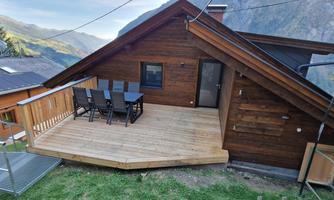 A wooden terrace with a table and chairs invites you to relax. In the background, green hills and mountains are visible.